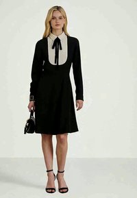 Mulher vestindo um vestido preto com gola branca e gravata-borboleta preta, segurando uma bolsa preta e usando saltos com tira no tornozelo pretos.