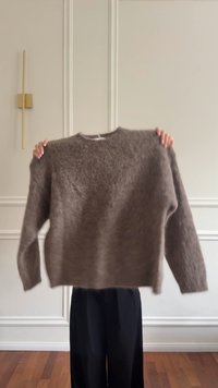 Brauner, flauschiger Pullover mit rundem Ausschnitt, langen Ärmeln und lockerer Passform, vor neutralem Hintergrund gehalten. Weiche Textur sichtbar.