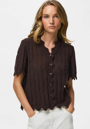 Jeune femme aux cheveux blonds portant un haut marron foncé texturé à manches courtes avec boutons et un pantalon blanc, les mains dans les poches, regardant devant elle.