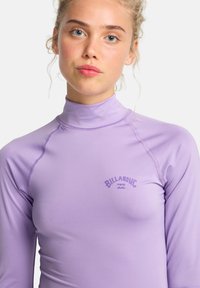 Rash guard di colore viola chiaro con maniche lunghe, colletto alto, cuciture piatte e un piccolo logo in viola scuro nella zona del petto.
