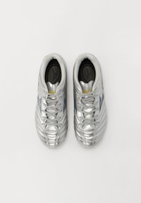 Scarpe da ginnastica argentate con una texture liscia e lucida, caratterizzate da un design con lacci, scanalature distintive e una soletta nera con un logo.