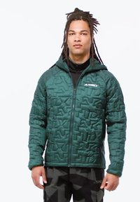 Groene geïsoleerde jas met een geometrisch quilted textuur, voorrits, capuchon en logo op de linkerkant van de borst. Gepaarde met donkere gestreepte broeken.