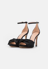 Melnas augstpapēžu sandales ar mezgla priekšpusi, potītes siksniņu, atvērtu pirkstu un satīna apdari. Iezīmē paaugstināta platforma un slaida papēža.