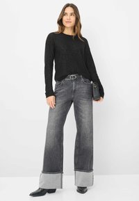Schwarzer Strickpullover mit strukturierter Oberfläche, kombiniert mit grauen, weit geschnittenen Jeans mit rohen Säumen und einem schwarzen Gürtel. Abgerundet wird der Look durch spitze schwarze Stiefel.