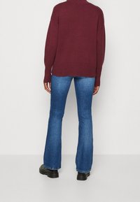 Pull en maille bordeaux surdimensionné avec un col montant, un ourlet côtelé et des épaules tombantes, associé à un jean en denim bleu évasé et des bottes noires.