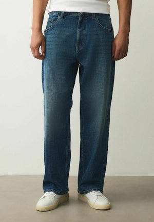 Mann mit blauen Jeans mit geradem Bein und weißen Sneakers, der vor einem neutralen Hintergrund steht.