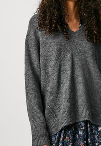 Grauer Wollpullover mit V-Ausschnitt und geripptem Saum. Er hat lange Ärmel und einen lässigen Schnitt, mit einer strukturierten, weichen Optik.