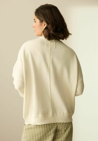 Cremefarvet sweatshirt med en afslappet pasform, der har en firkantet stikning detaljeret ved nakken bagpå og ribbet kant. Sammen med ternede bukser.