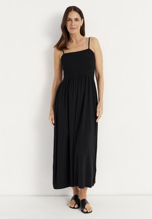 Frau mit schulterlangem braunem Haar, die ein ärmelloses schwarzes Maxikleid und schwarze Sandalen trägt, vor weißem Hintergrund stehend.