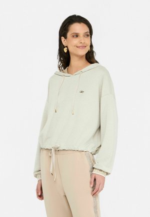 Felpa con cappuccio grigio chiaro, con orlo elasticizzato e coulisse; presenta dettagli dorati e una texture sottile. Abbinata a pantaloni beige.