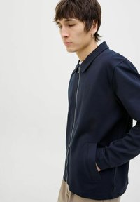 Jūrinės mėlynos zip-up striukės, pagamintos iš lygaus audinio su apykakle, šoninėmis kišenėmis ir subtiliu blizgesiu. Fituotas siluetas su tiesiu kirpimu.