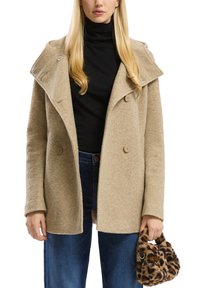 Bež tekstuuriga mantel laia krae, topelt nööpide ja lõdva kroogiga, kantud musta kõrge kaelusega särk üle. Hoiab leopardimustriga käekotti.