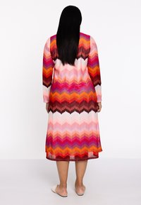 Maxikleid mit Chevron-Muster in lebhaften Rosa-, Orange- und Rottönen. Lange Ärmel, gerippte Textur und lockerer Schnitt.