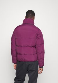 Kurtka puchowa w głębokim fiolecie z wysokim kołnierzem, pikowanym wzorem i elastycznymi mankietami. Charakteryzuje się gładką fakturą i minimalistycznym stylem.