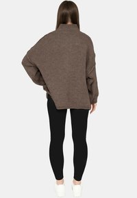 Vrouw met lang bruin haar die een losse, bruin gebreide trui draagt, zwarte legging en witte sneakers, gezien van achteren tegen een effen achtergrond.