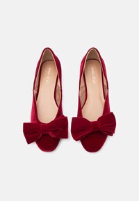 Κόκκινες βελούδινες μπαλαρίνες με μεγάλο φιόγκο στο μπροστινό μέρος, στρογγυλή μύτη και λεία εσωτερική επένδυση, διαθέτοντας λογότυπο στο εσωτερικό.