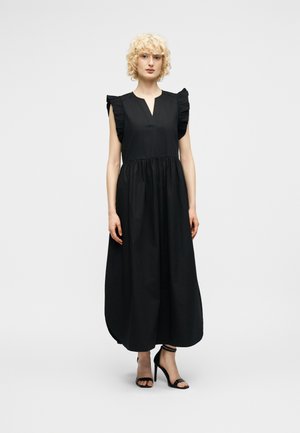 Kvinde med kort krøllet blondt hår iført en sort ærmeløs midi-kjole med flæser på skuldrene og sorte højhælede sandaler, stående.