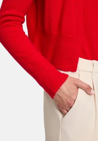 Cardigan en tricot rouge avec des poignets côtelés assorti à un pantalon crémeux sur mesure. Main placée dans la poche, illustrant un design et un ajustement décontractés.