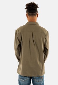 Camisa verde oliva con pliegue en la espalda, cuello tradicional, mangas largas y un corte relajado. Combinada con jeans de mezclilla azules.
