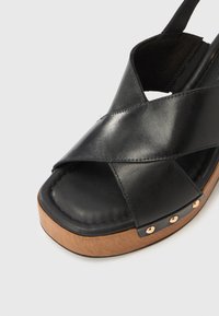 Μαύρο δερμάτινο σανδάλι με σχεδιασμό σταυρωτού λουριού, ξύλινη πλατφόρμα στην σόλα και διακοσμητικά μεταλλικά καρφάκια στο πλάι. Στυλ με ανοιχτά δάχτυλα.