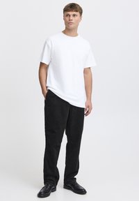 Maglietta bianca a girocollo, abbinata a pantaloni neri in velluto a coste. Il modello è in piedi, indossa scarpe nere, design minimalista, tessuto dalla texture liscia.