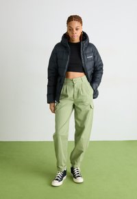 Svart pufferjakke med hette, iført en crop top og lysegrønne cargo-bukser. Svart høyhælte sneakers med hvite såler.