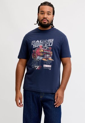 Mand med flettet hår og skæg iført en mørkeblå T-shirt med en rød racerbilsillustration og fart-tema tekst, stående foran en ensfarvet baggrund.