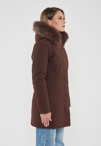 Parka marrone con cappuccio foderato in pelliccia sintetica, texture liscia, tasche laterali e silhouette aderente, indossato sopra jeans azzurri chiari.