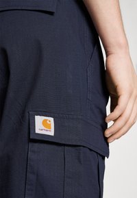 Marineblå cargo bukser laget av teksturert stoff. Har en stor lomme med et Carhartt-logo merket. Nærbilde av livlinjen og lomområdet.
