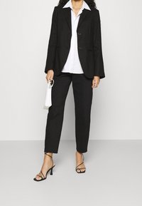 Blazer structuré noir à deux boutons, associé à une chemise blanche à col et un pantalon noir fuselé. Accompagné de sandales à talons noires à lanières.