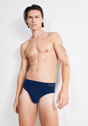 Skjorteløs mand med mellemlangt brunt hår iført marineblå Sloggi underbukser, står foran en hvid baggrund.