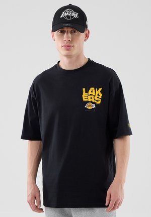 Joven con gorra negra de Los Angeles Lakers y camiseta negra con el logo amarillo de los Lakers en el pecho, de pie frente a un fondo gris.