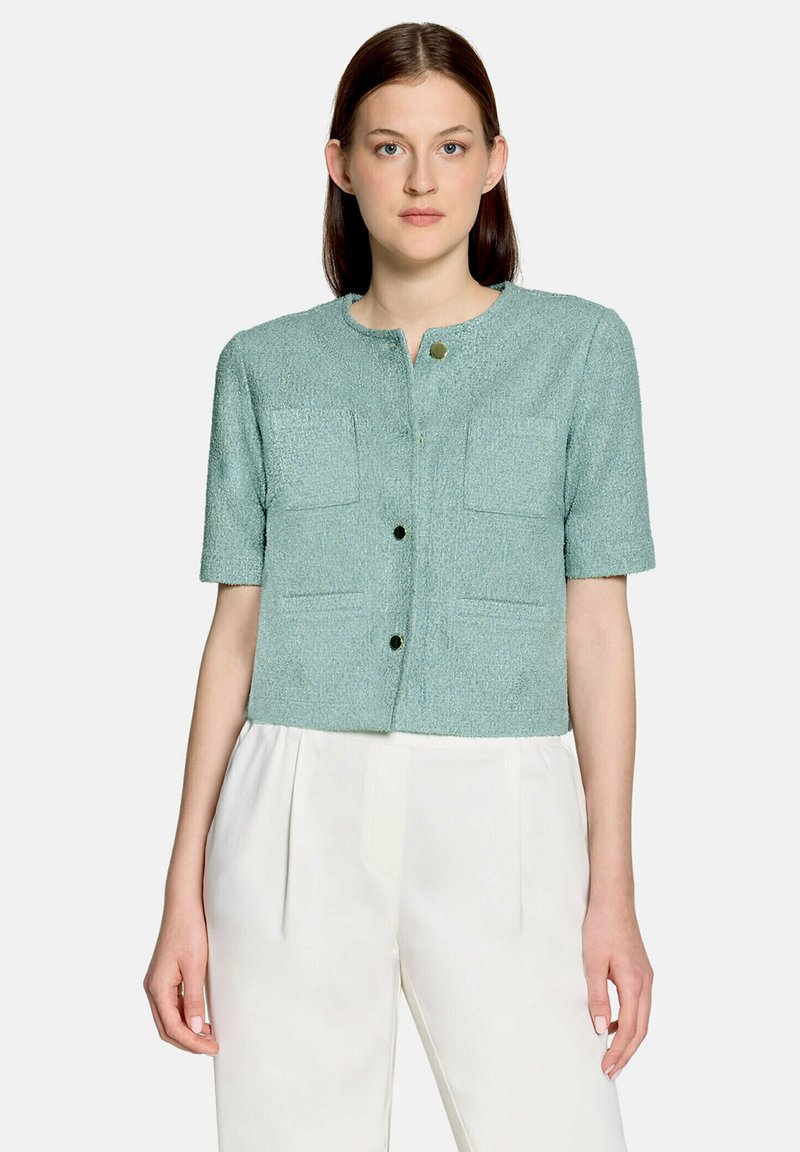 Femme portant une veste texturée à manches courtes bleu clair avec des boutons dorés et un pantalon blanc plissé taille haute.