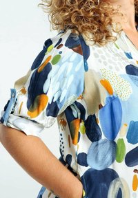Personne aux cheveux bouclés portant une chemise à manches longues avec des motifs abstraits colorés en bleu, orange, vert et blanc, bras plié à la taille.