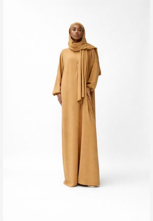 Kobieta ubrana w luźną, długą suknię w kolorze wielbłądziej wełny z pasującym hidżabem, stojąca na jednolitym białym tle.