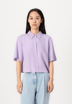 Short-sleeve, cropped shirt in light lavender, featuring a button-up front, pointed collar, and smooth fabric. Paired with light denim trousers.
