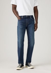 Jeans in denim blu scuro con taglio dritto, design standard a cinque tasche e hardware con bottoni metallici. Indossati con sneakers nere e bianche.