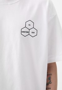 White cotton t-shirt featuring a black hexagonal graphic with the text "ALL CONDITIONS GEAR." Short sleeves and a crew neckline.