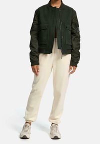 Groene bomberjack met een wollen lichaam en satijnen mouwen, voorzien van twee voorzakken en een ritssluiting, gecombineerd met crèmekleurige joggers en sneakers.