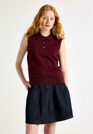Ung kvinde med langt rødt hår iført en ærmeløs bordeaux polo og en mørkeblå denim nederdel, stående med hænderne i lommerne.
