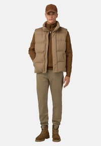 Hnědá péřová vesta přes hnědý pletený svetr, doplněná světle hnědými kalhotami a hnědými šněrovacími botami, které mají strukturovaný pletený vzor.