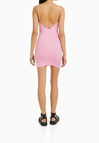 Roze bodycon jurk met dunne bandjes en gekruiste rugdetail. Glad textuur, knielange snit. Gecombineerd met zwarte met studs versierde sandalen.