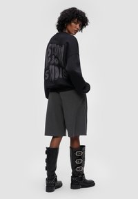 Felpa nera oversize con testo grigio stilizzato sul retro, abbinata a pantaloni corti grigi su misura e stivali neri alti fino al ginocchio con fibbie.