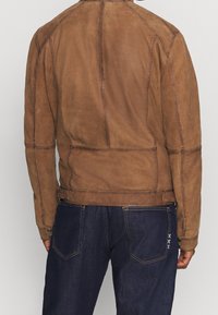 Vista posteriore di una persona che indossa una giacca in suede marrone con cuciture dettagliate e jeans blu scuro con ricamo bianco a croce su una tasca.