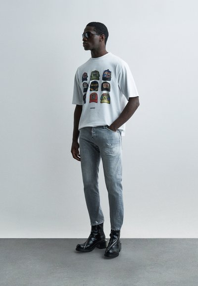 Άνδρας που φορά μαύρα γυαλιά ηλίου, λευκό μπλουζάκι με πολύχρωμα γραφικά καπέλου, ανοιχτό μπλε τζιν και μαύρα δερμάτινα μποτάκια, στέκεται μπροστά σε ένα απλό φόντο.