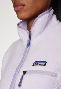 Lys lavendel fleecejakke med høy krage, halv glidelås og frontlomme. Har en svart Patagonia-logo og teksturert stoff.