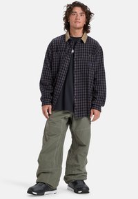 Schwarz-lila karierter Button-Up-Hemd mit beigem Kragen, über einem schwarzen Hemd getragen; kombiniert mit lockeren olivgrünen Hosen und schwarzen Schuhen.