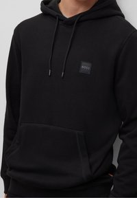 Zwarte hoodie met een voorzak, een capuchon met trekkoord en een klein zwart logo-patch. Zacht materiaal met een gladde textuur en geribde manchetten.