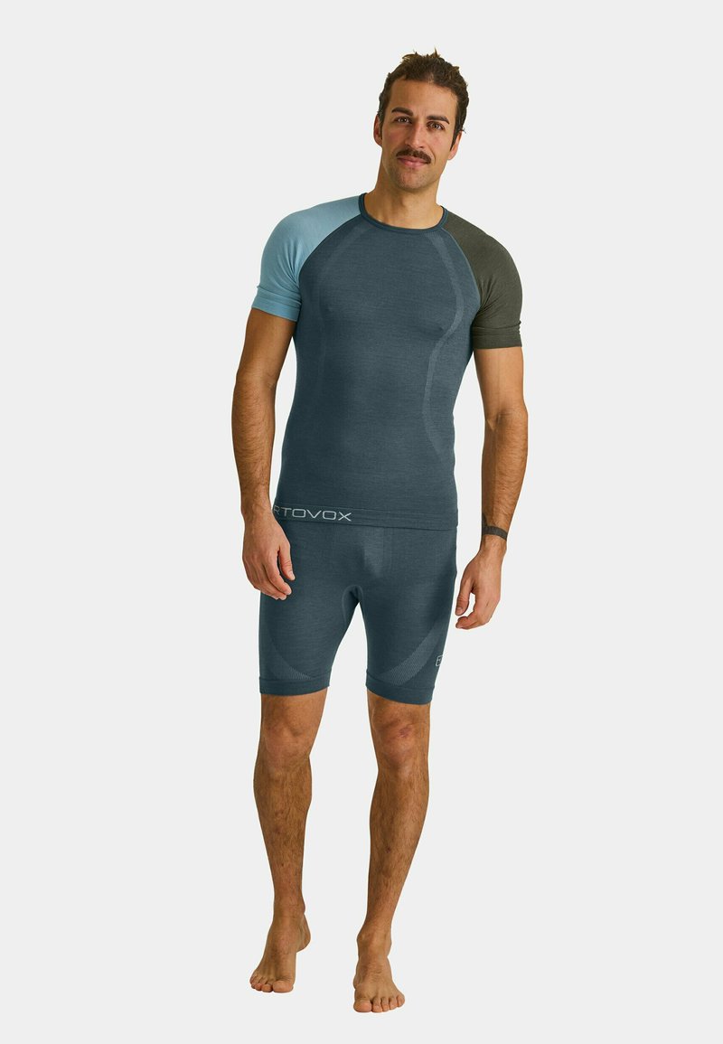 Man with mustache wearing a fitted short-sleeve shirt with differently colored sleeves and matching shorts, standing barefoot against white background.