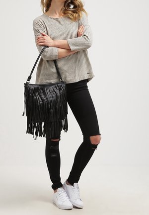 Sac à main en cuir noir avec franges et bandoulière, porté avec un haut rayé à manches longues, un jean noir déchiré et des baskets blanches.